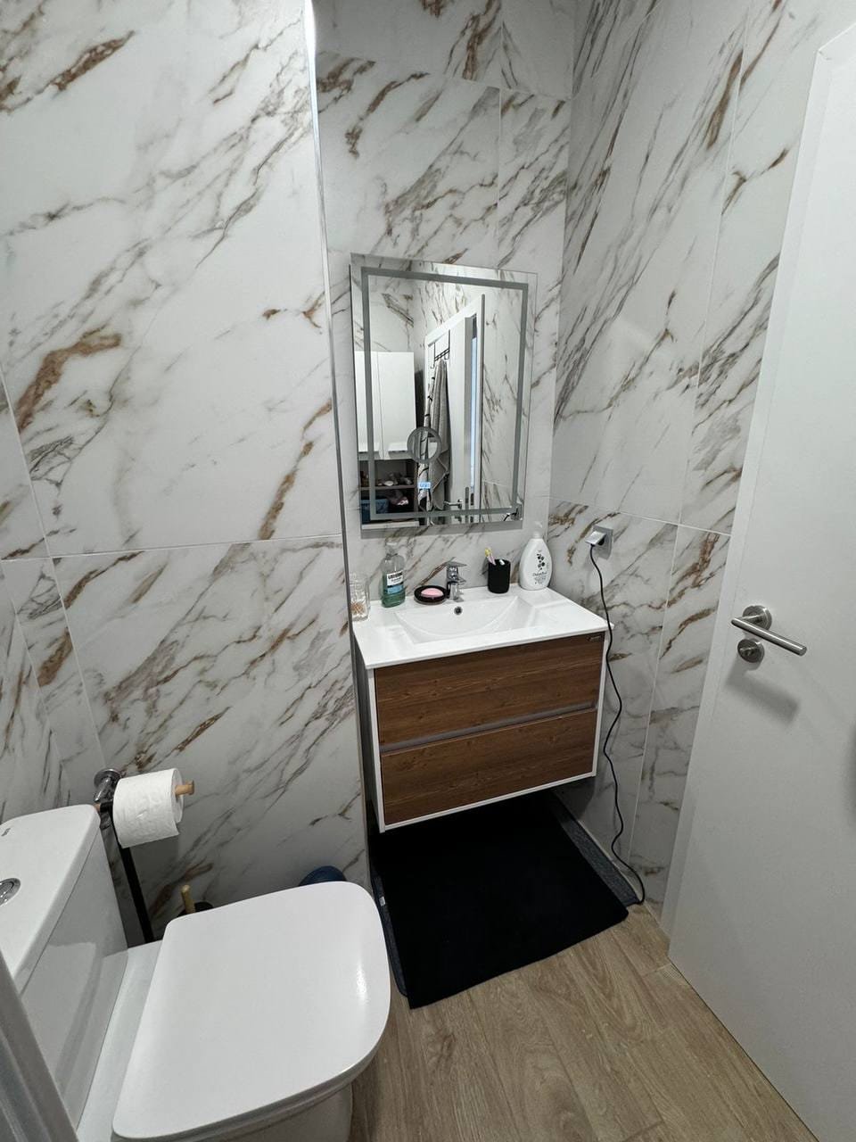 Современный санузел с мраморными стенами, раковиной на деревянной тумбе и зеркалом, удобный для жизни в Болгарии.