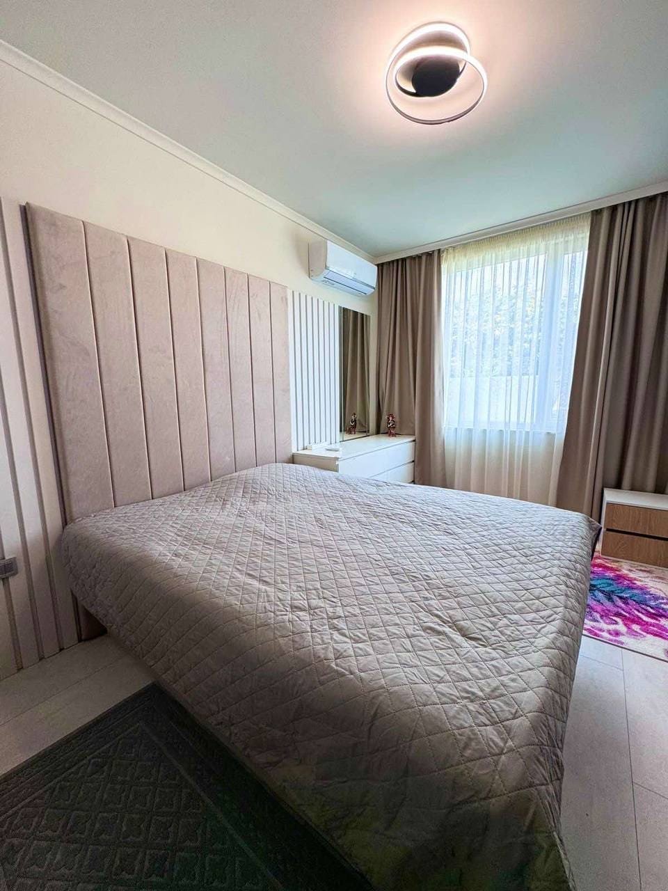 Современная спальня с большой кроватью, светлыми шторами и комфортной атмосферой, идеальная для отдыха в Болгарии.