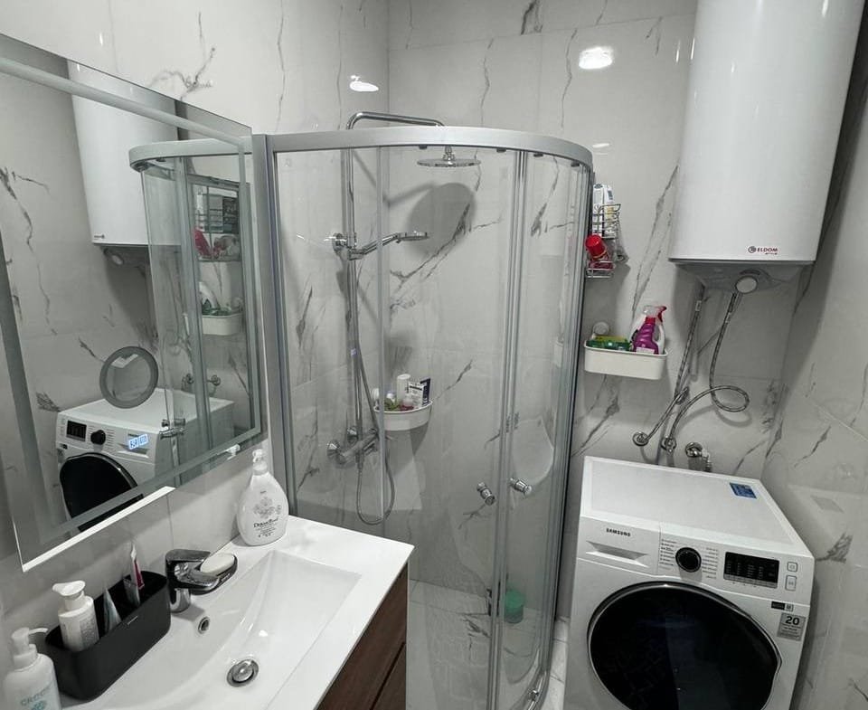 Современная ванная комната с душевой кабиной, стиральной машиной и зеркалом, оформленная в нейтральных тонах.