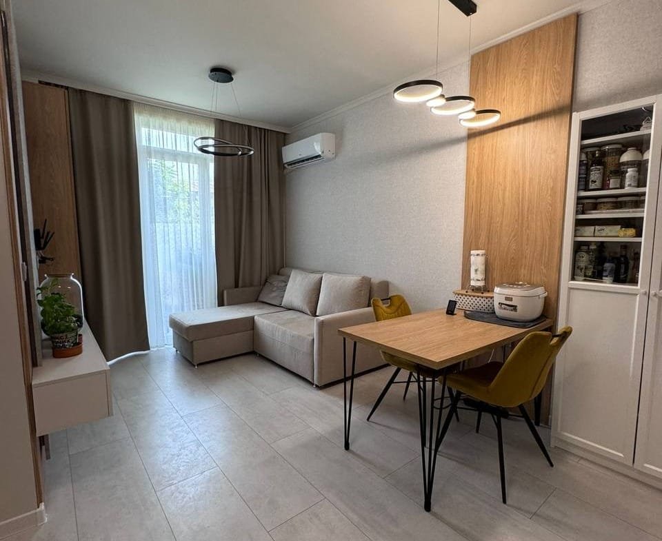 Современный светлый интерьер квартиры в Болгарии с уютной зоной отдыха, столом для еды и кухонными принадлежностями.