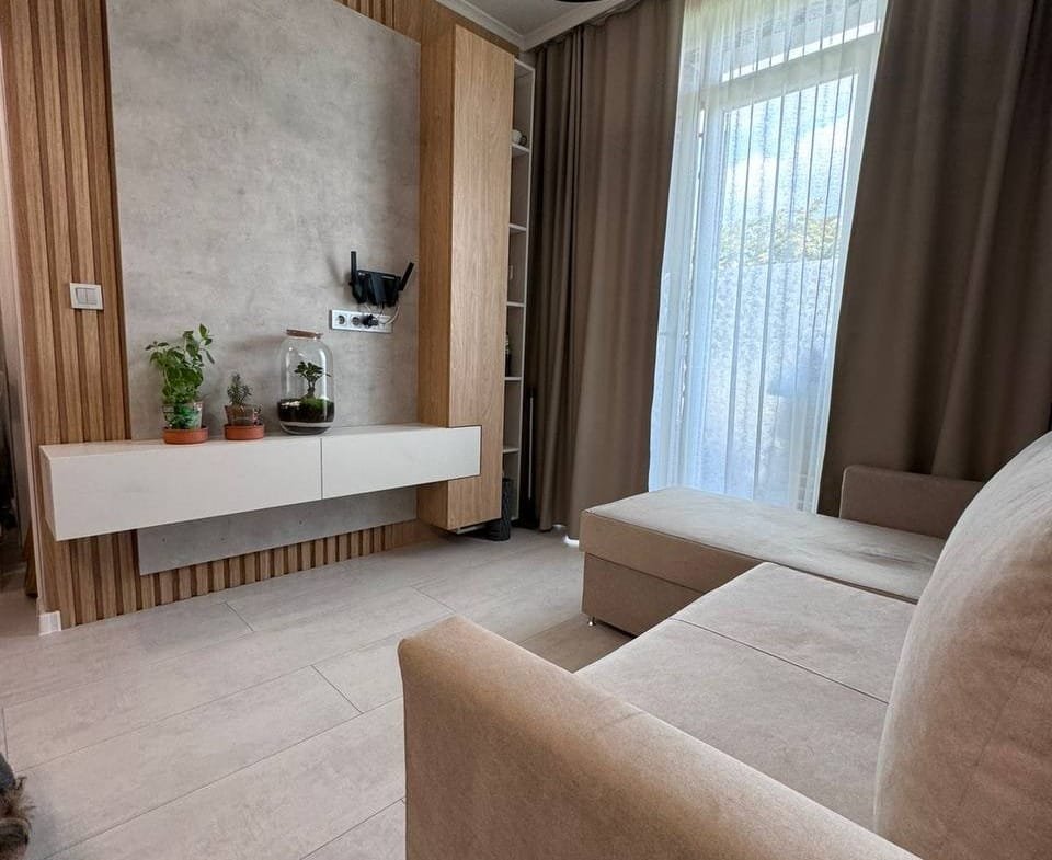 Современная гостиная с уютным диваном, деревянными элементами, освещением и большим окном, идеально подходящая для продажи недвижимости в Болгарии.