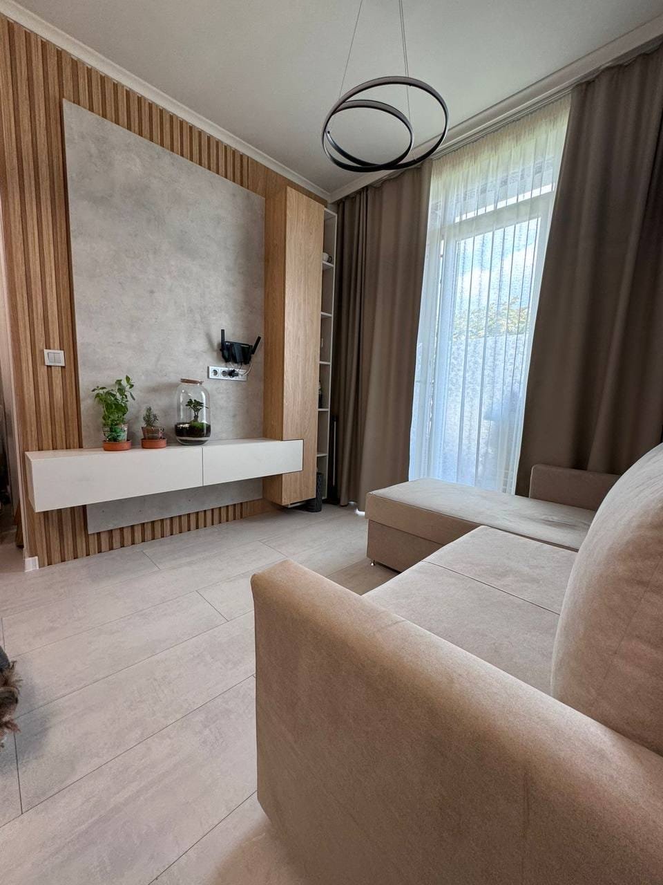 Современная гостиная с уютным диваном, деревянными элементами, освещением и большим окном, идеально подходящая для продажи недвижимости в Болгарии.