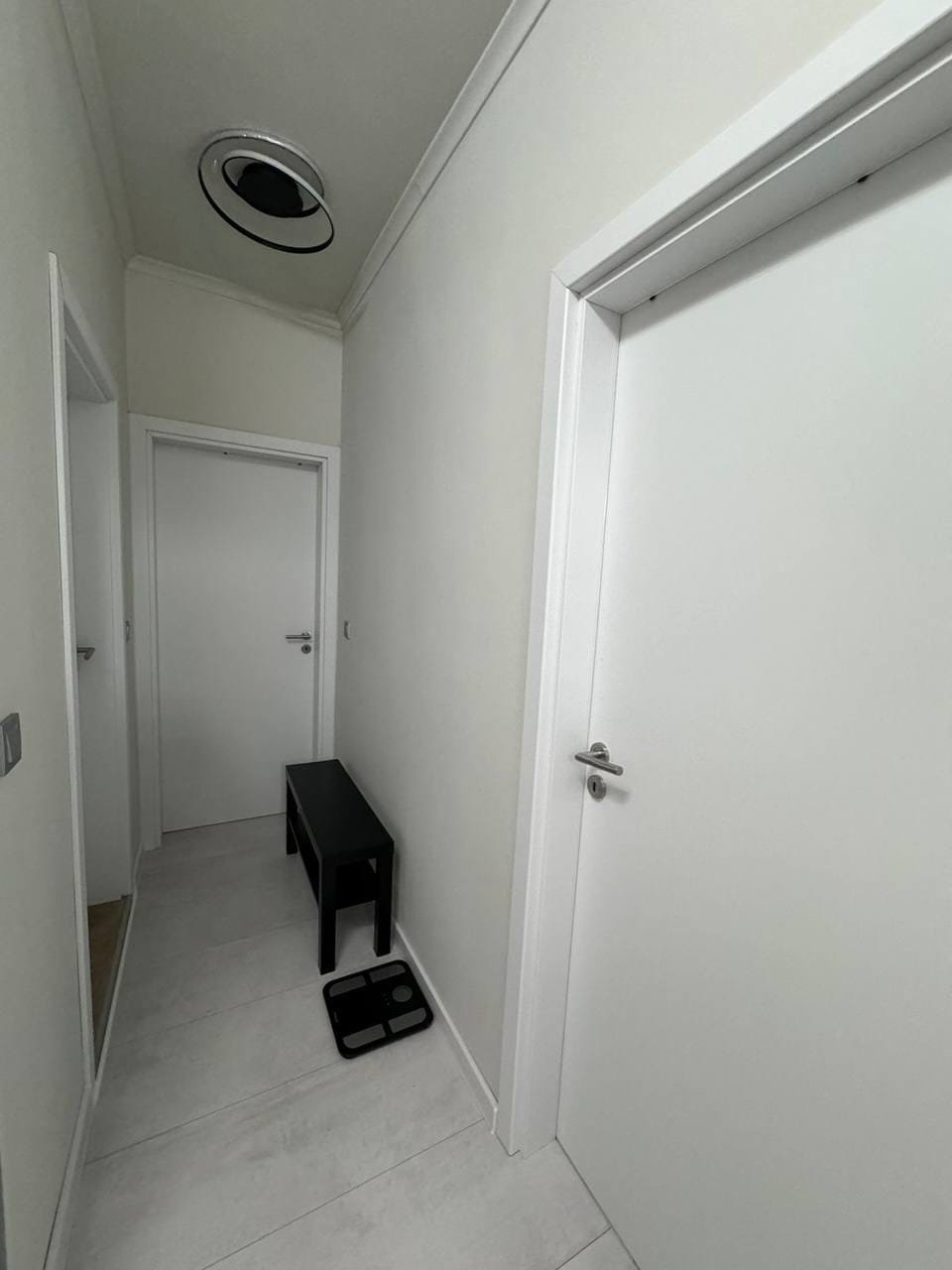 Современный коридор с белыми стенами, дверями и черной табуреткой, демонстрирующий спокойный интерьер для покупки недвижимости в Болгарии.