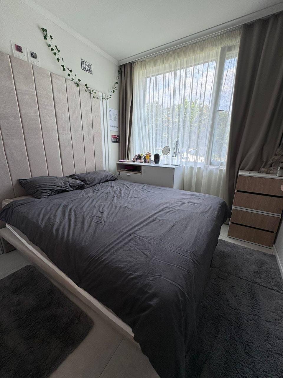 Современная спальня с серым постельным бельем, большой окном и минималистичной мебелью, идеальная для комфортного проживания в Болгарии.