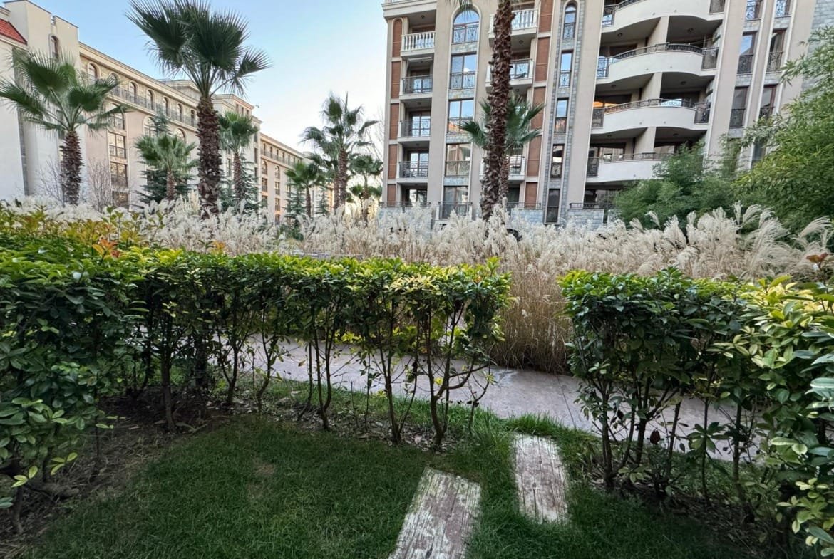 Зеленый сад с кустами и пальмами на фоне жилого комплекса в Болгарии, идеальное место для жизни и инвестиций в недвижимость.