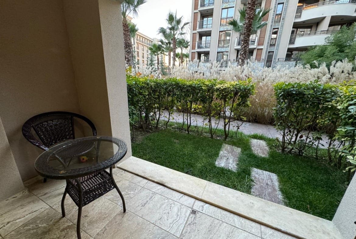 Балкон с креслом и столиком, зеленый газон и кусты, вид на жилой комплекс в Болгарии, идеальное место для отдыха.