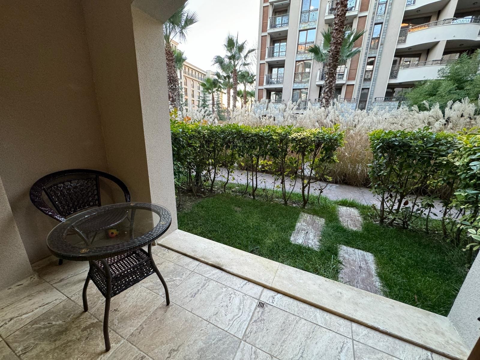 Балкон с креслом и столиком, зеленый газон и кусты, вид на жилой комплекс в Болгарии, идеальное место для отдыха.