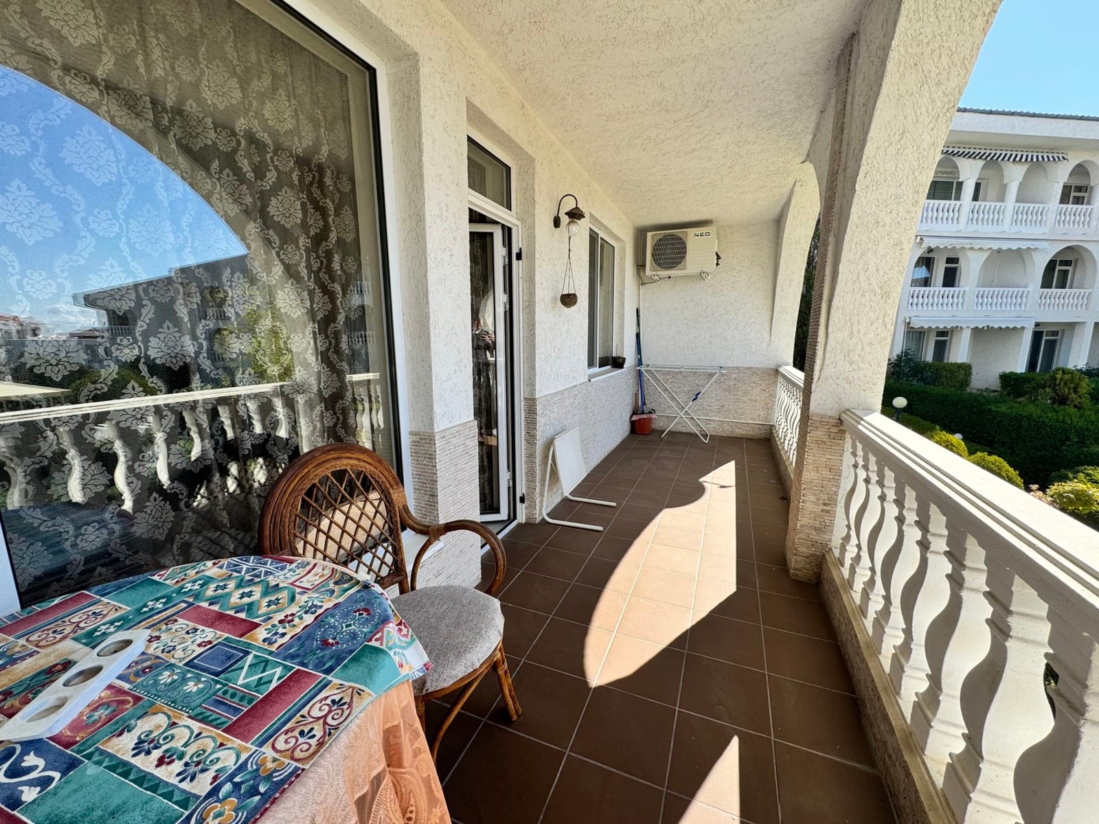 Широкая веранда с плетёной мебелью, столом и сушилкой, окружённая зелеными насаждениями в Болгарии.