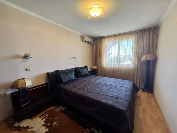 Современная спальня с большой двуспальной кроватью, светлыми шторами и мягким освещением, идеально подходит для отдыха в Болгарии.