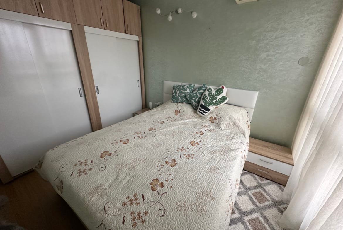 Спальня с двуспальной кроватью, украшенной цветочным покрывалом, и стильным ночным столиком рядом. Теплые оттенки интерьера.