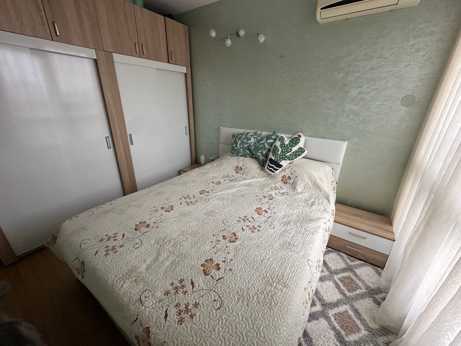 Спальня с двуспальной кроватью, украшенной цветочным покрывалом, и стильным ночным столиком рядом. Теплые оттенки интерьера.