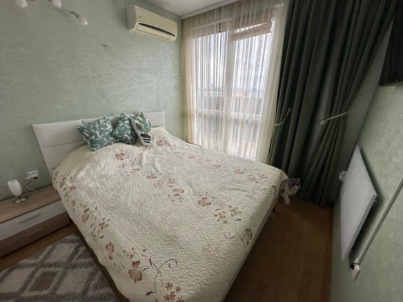 Уютная спальня с двуспальной кроватью, зелеными шторами, подушками с цветочным узором и ковром на деревянном полу.