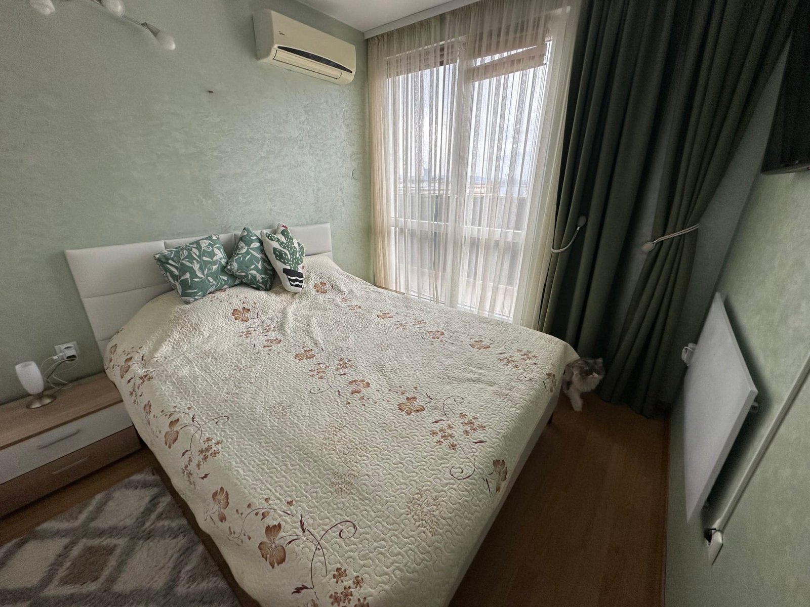 Уютная спальня с двуспальной кроватью, зелеными шторами, подушками с цветочным узором и ковром на деревянном полу.