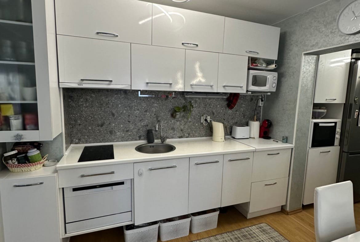 Современная кухня с белыми шкафами, серой стеновой плиткой и светлым деревянным полом. Освещение в виде кольца, стол и стулья.