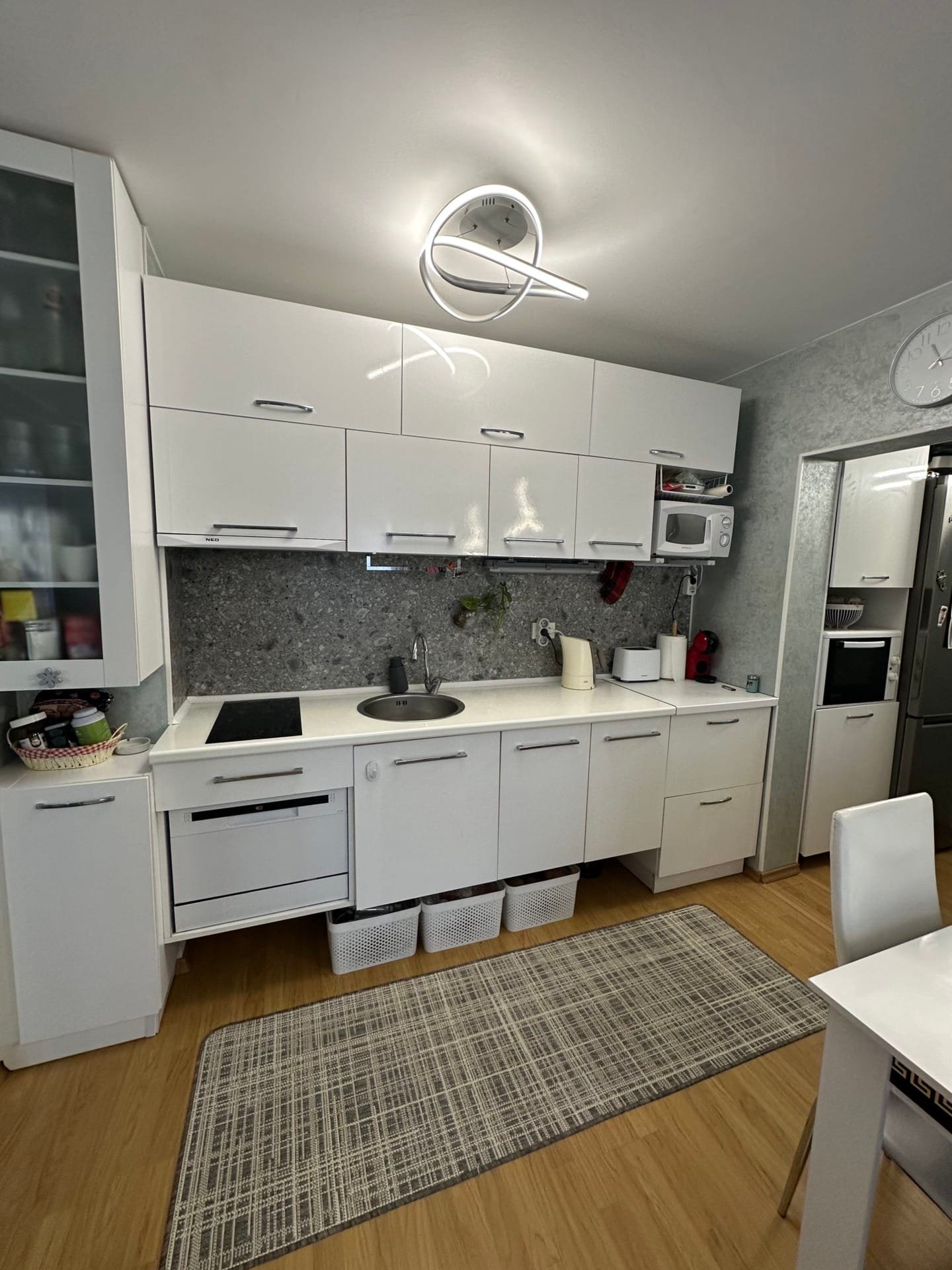 Современная кухня с белыми шкафами, серой стеновой плиткой и светлым деревянным полом. Освещение в виде кольца, стол и стулья.