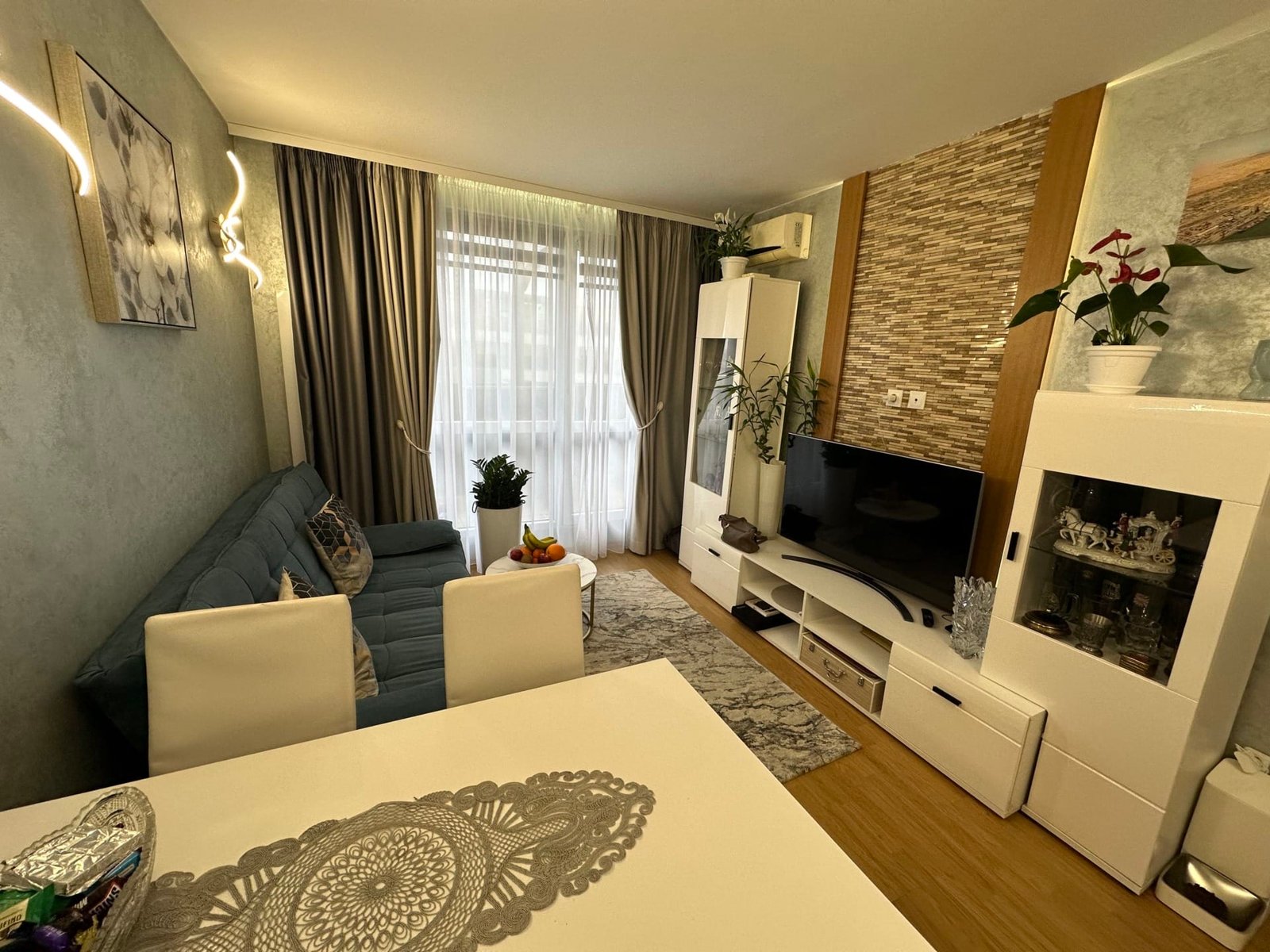 Современный интерьер гостиної с голубым диваном, белыми мебелью и декоративными растениями, гармоничный и светлый.