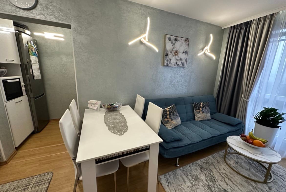 Современная гостиная с синим диваном, белым столом, стульями и кухонным оборудованием, оформленная в светлых тонах.