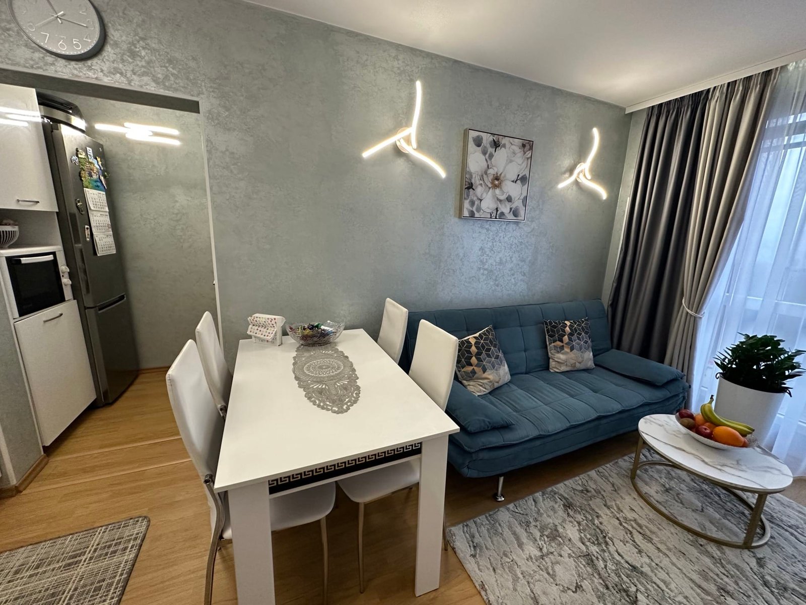 Современная гостиная с синим диваном, белым столом, стульями и кухонным оборудованием, оформленная в светлых тонах.