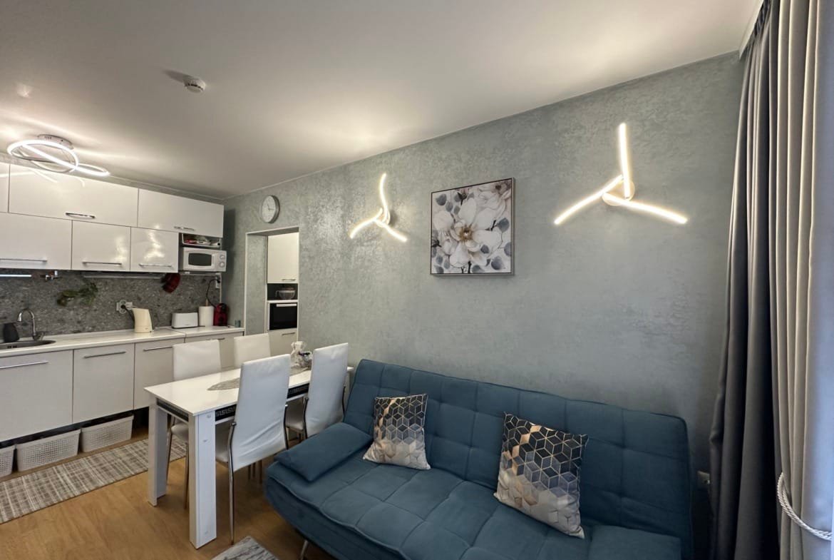 Современный интерьер кухни-студии с синим диваном, светлыми стенами и стильным освещением. Стол с четырьмя стульями.