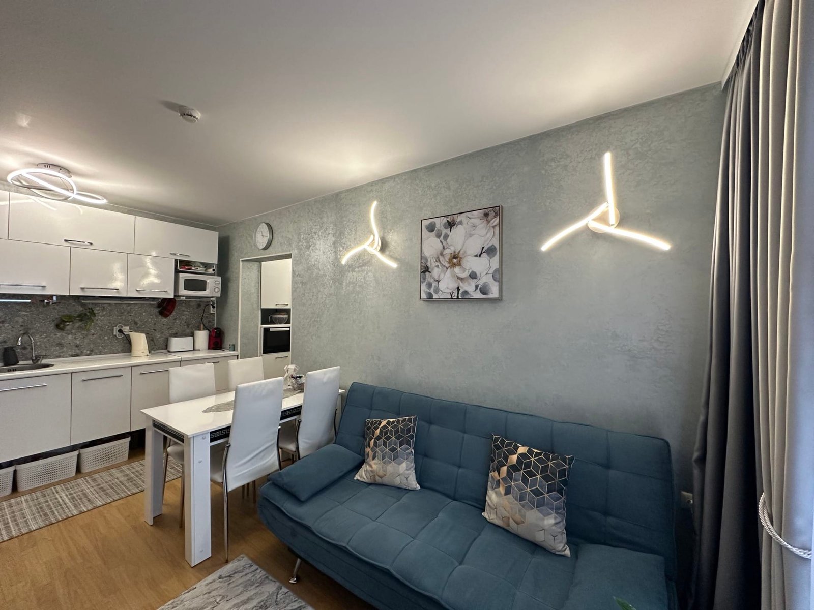 Современный интерьер кухни-студии с синим диваном, светлыми стенами и стильным освещением. Стол с четырьмя стульями.