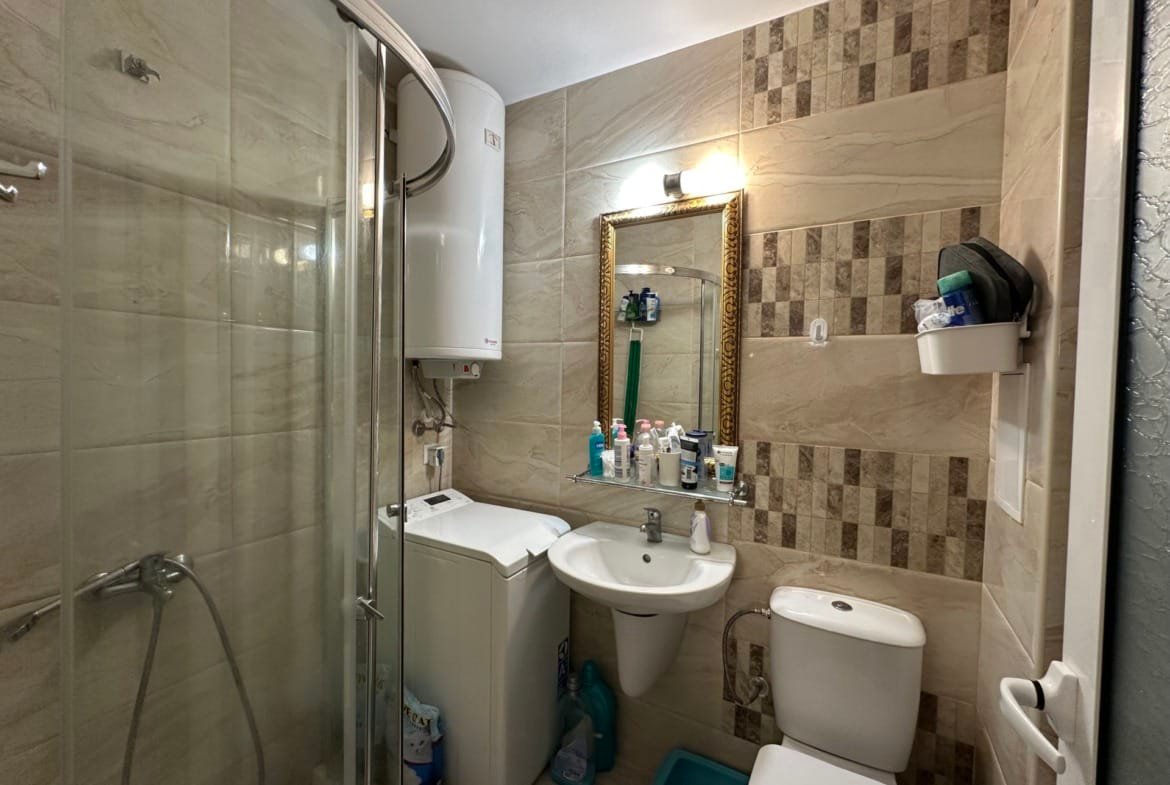 Санузел с душевой кабиной, зеркалом в золотой рамке, умывальником, стиральной машиной и полками с косметикой.