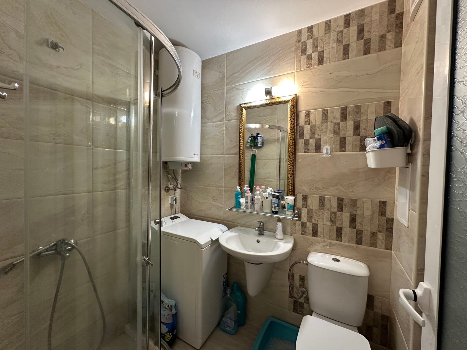 Санузел с душевой кабиной, зеркалом в золотой рамке, умывальником, стиральной машиной и полками с косметикой.