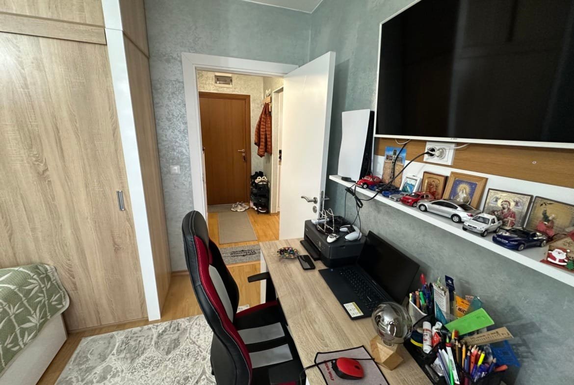 Современный офисный уголок с рабочим столом, креслом, компьютером и полкой с автомобилями на стене.