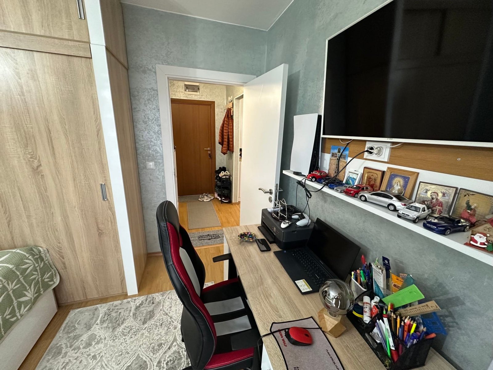 Современный офисный уголок с рабочим столом, креслом, компьютером и полкой с автомобилями на стене.