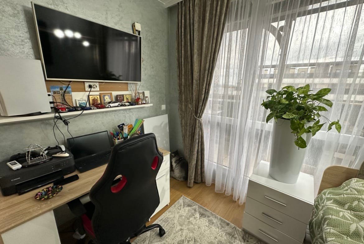 Современный домашний офис с письменным столом, компьютером, растением в горшке и светлыми занавесками.