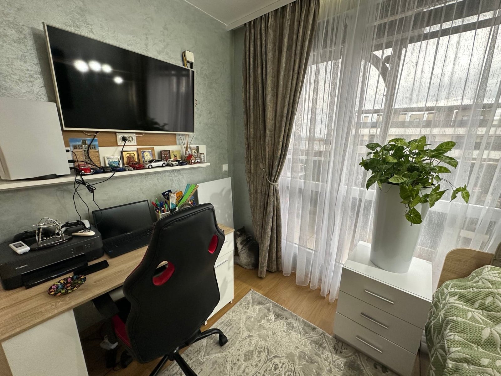 Современный домашний офис с письменным столом, компьютером, растением в горшке и светлыми занавесками.