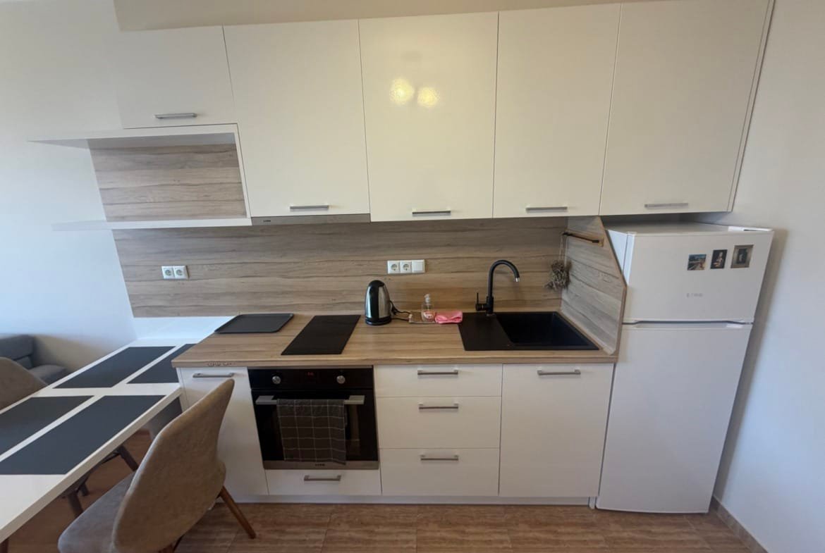 Современная кухня с белыми фасадами, деревянными акцентами, плитой, холодильником и обеденной зоной в квартире в Болгарии.