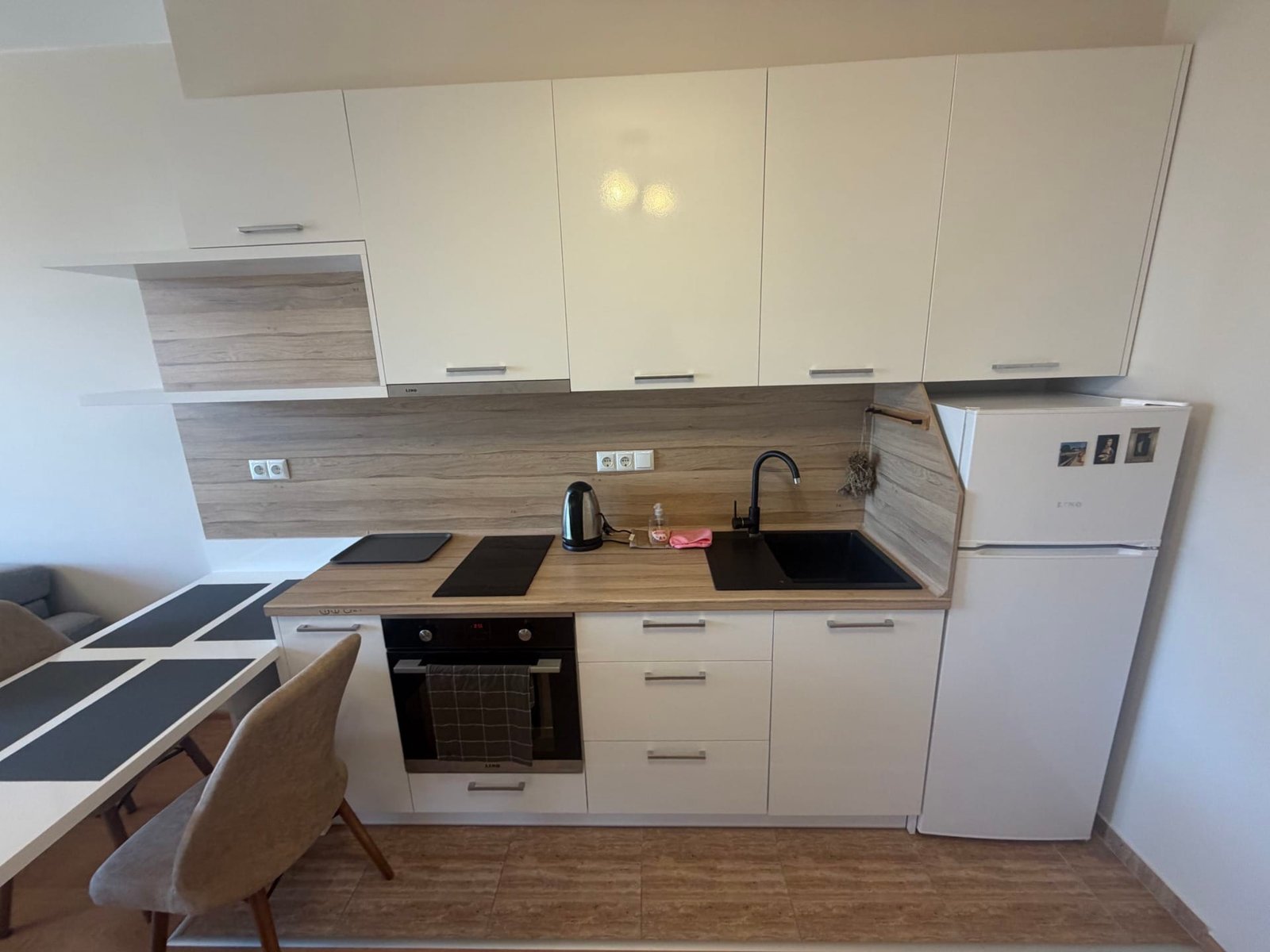 Современная кухня с белыми фасадами, деревянными акцентами, плитой, холодильником и обеденной зоной в квартире в Болгарии.