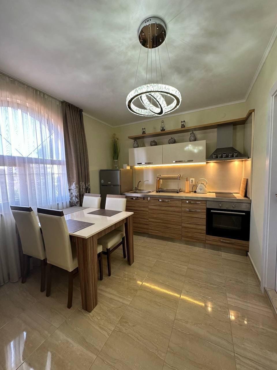 Современная кухня в светлых тонах с обеденной зоной, стильной люстрой и оборудованием для комфортного проживания в Болгарии.