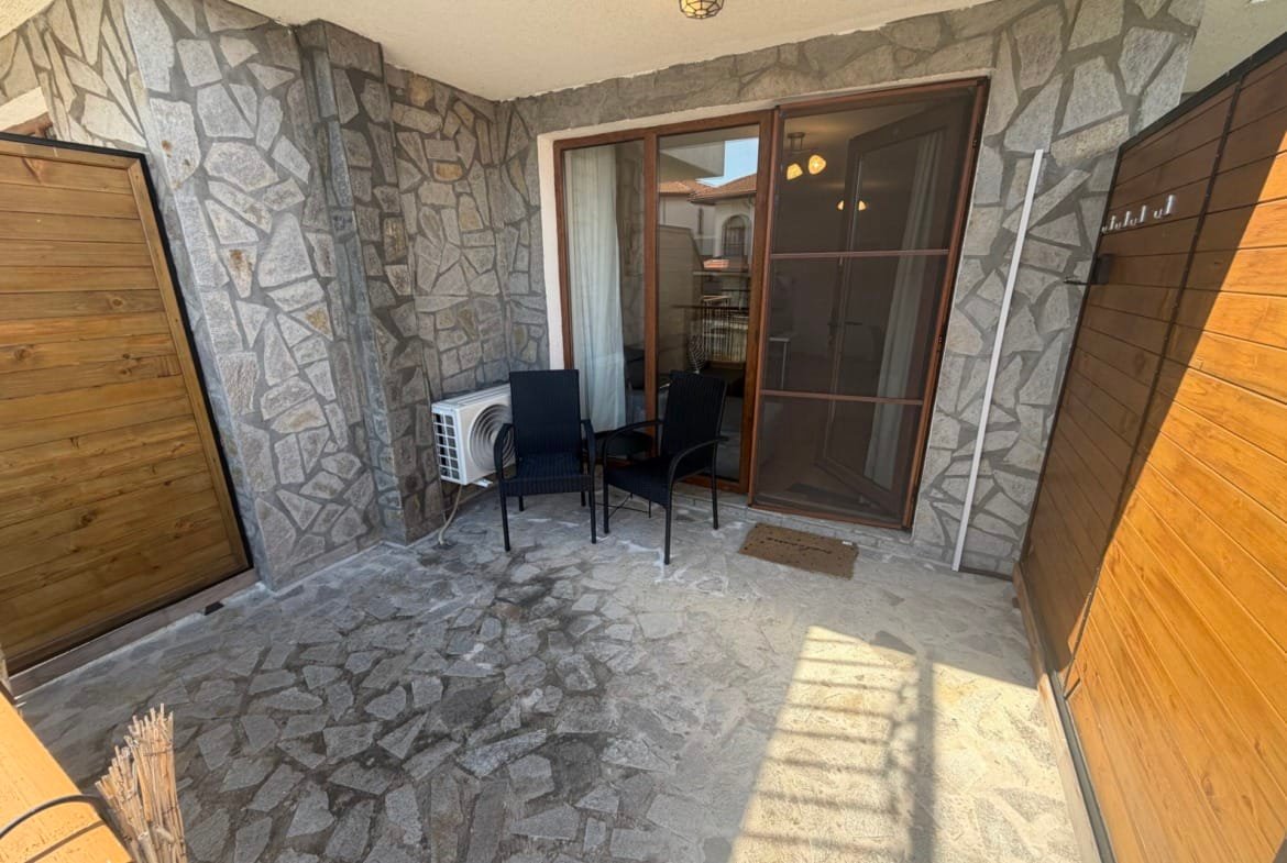 Уютная терраса с каменной отделкой, стальными стульями и выходом на зелёный двор в болгарском комплексе недвижимости.