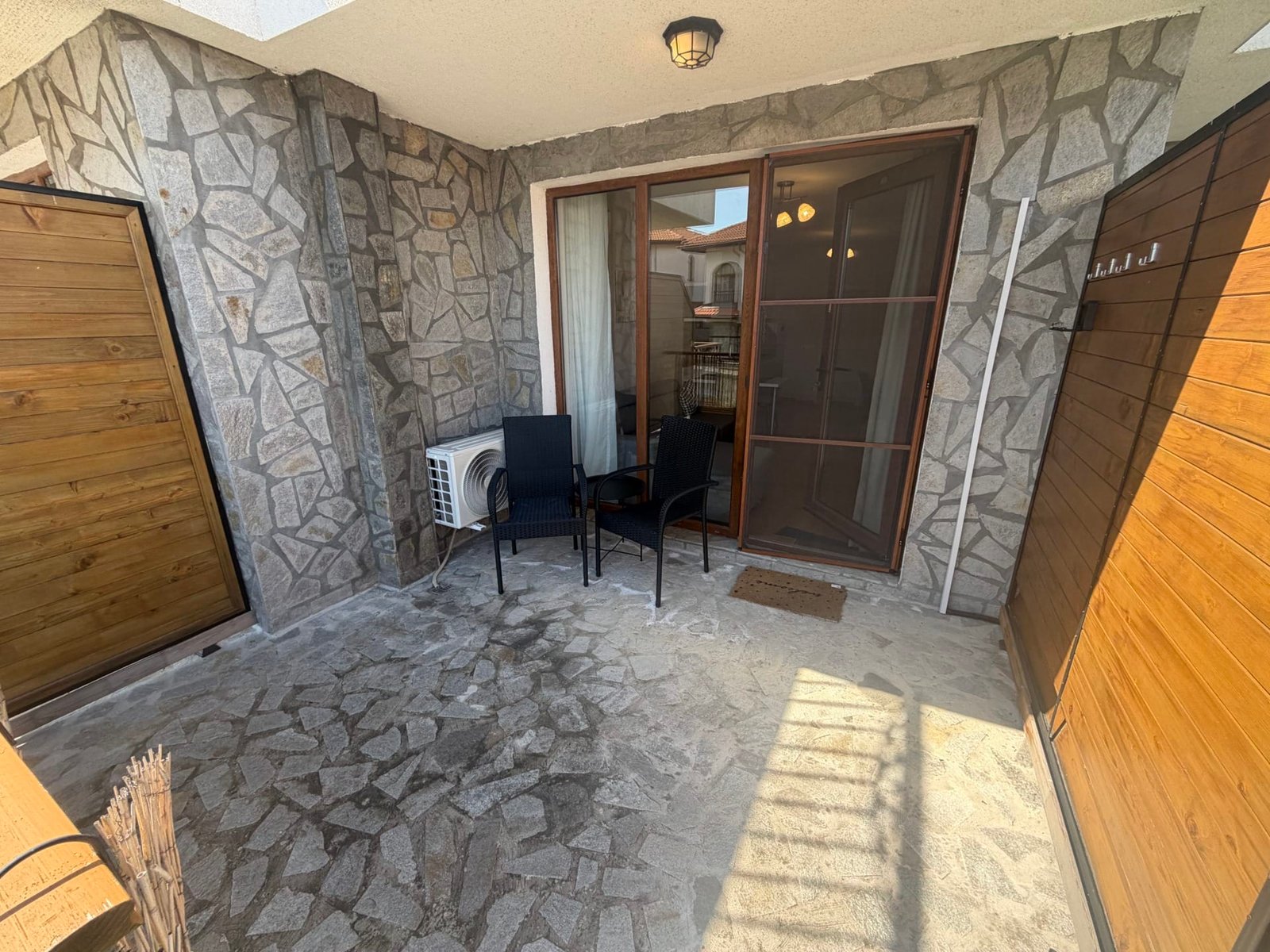 Уютная терраса с каменной отделкой, стальными стульями и выходом на зелёный двор в болгарском комплексе недвижимости.