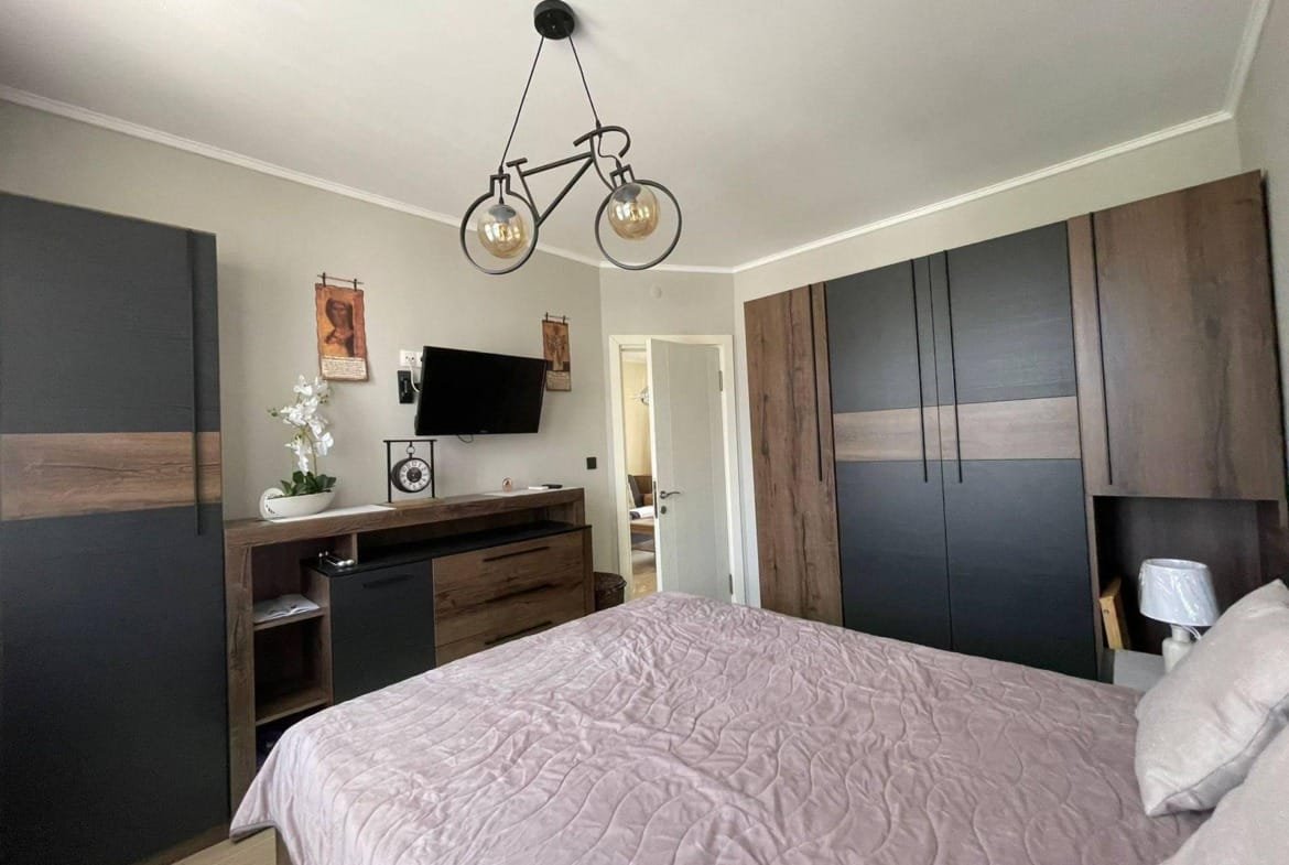 Современная спальня с двухспальной кроватью, шкафом и телевизором, оформленная в нейтральных тонах, подходящая для продажи недвижимости в Болгарии.