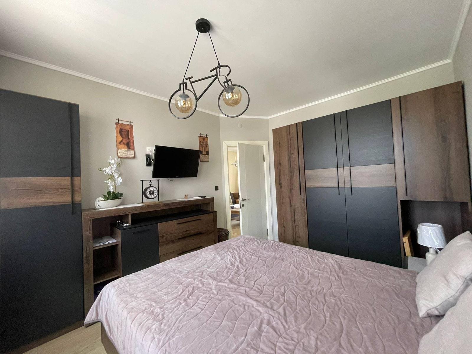 Современная спальня с двухспальной кроватью, шкафом и телевизором, оформленная в нейтральных тонах, подходящая для продажи недвижимости в Болгарии.