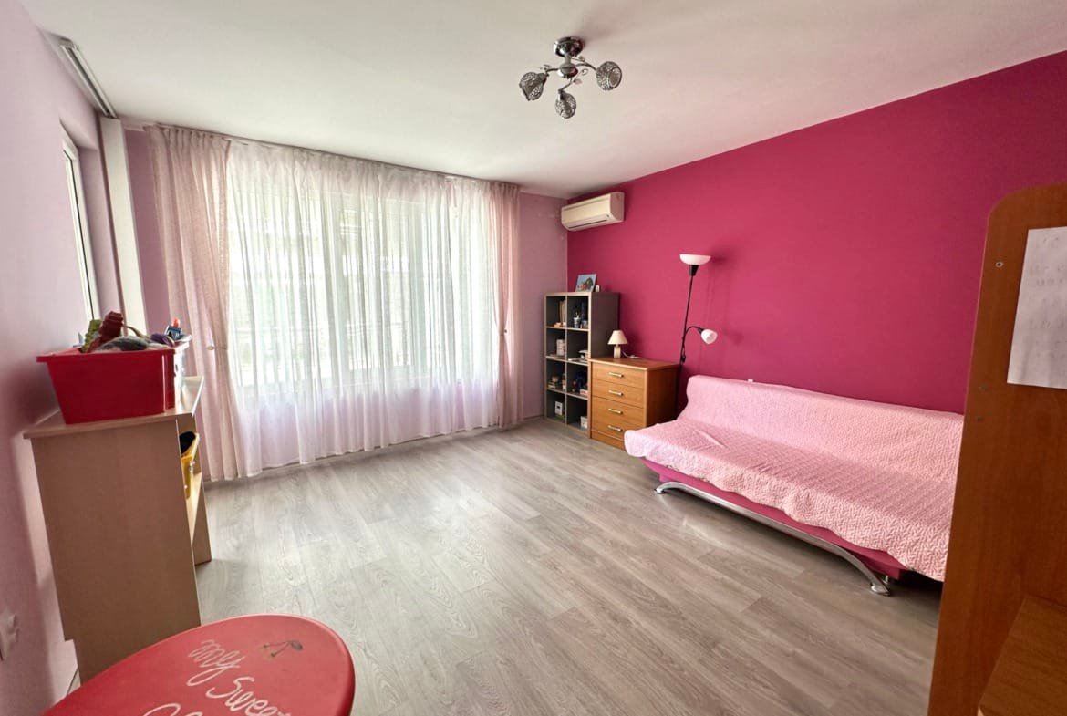 Просторная комната с розовыми стенами, окнами с занавесками, диваном и мебелью, подходящая для покупки недвижимости в Болгарии.