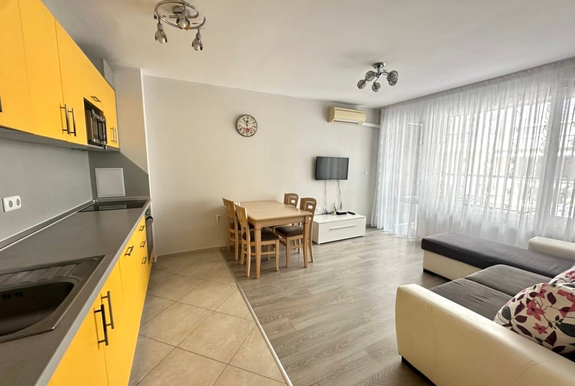 Светлая гостиная с кухней и обеденной зоной, желтые шкафы, деревянный стол и диван, идеальна для покупки недвижимости в Болгарии.