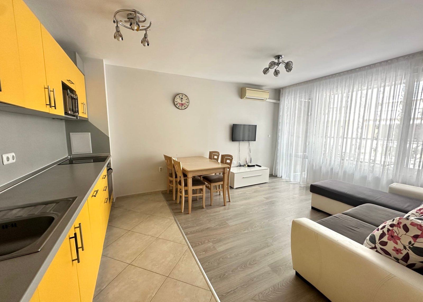 Светлая гостиная с кухней и обеденной зоной, желтые шкафы, деревянный стол и диван, идеальна для покупки недвижимости в Болгарии.