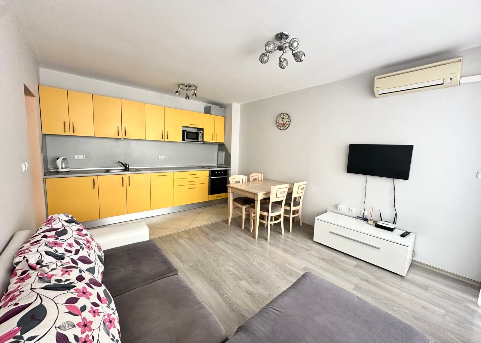 Современная квартира с кухней в жёлтых тонах, столом и стульями, удобным диваном и телевизором в Болгарии.