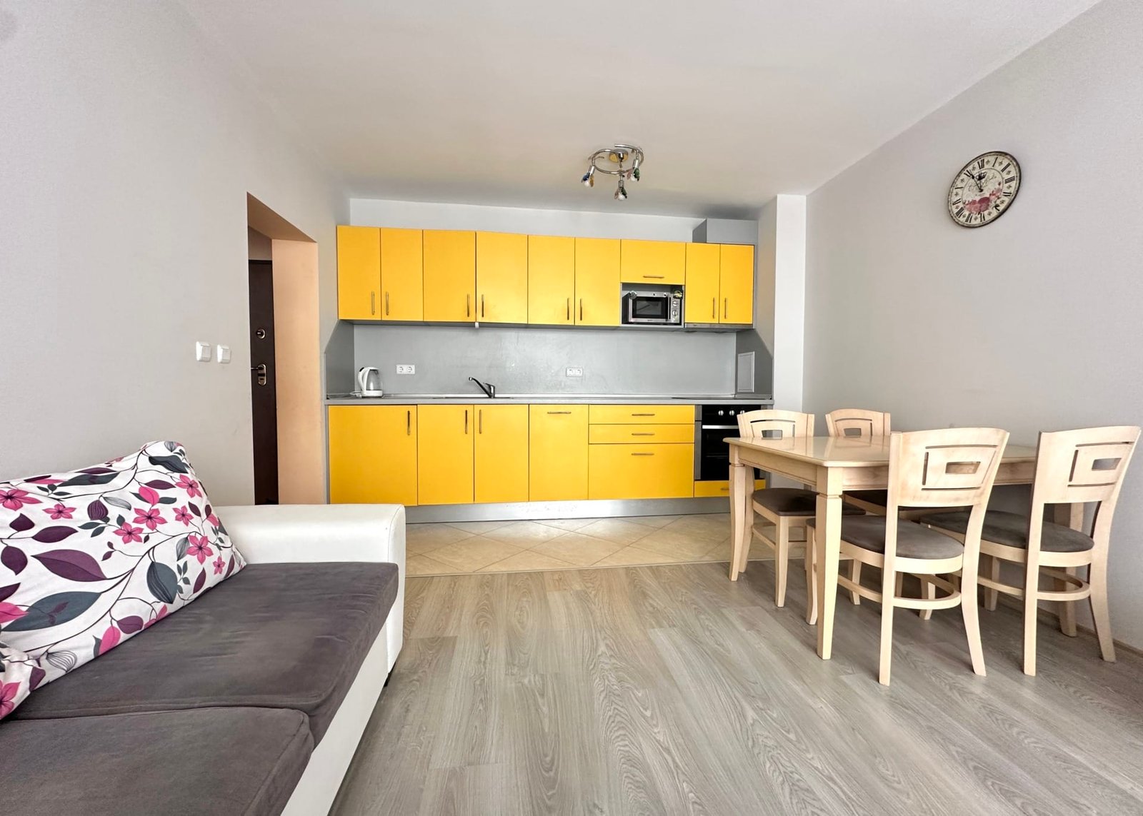 Кухня-гостиная с желтыми шкафами, столом и стульями, диваном и часами, подходит для продажи недвижимости в Болгарии.