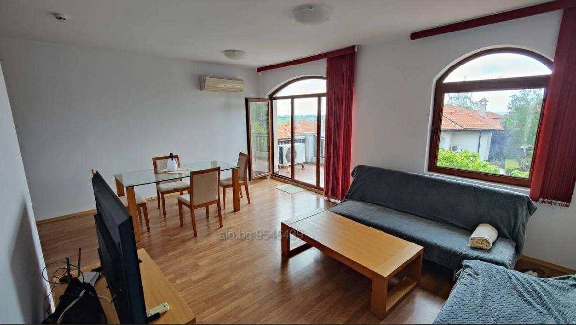 Светлая гостиная с большим окном, мебелью и выходом на балкон, идеальная для покупки недвижимости в Болгарии.