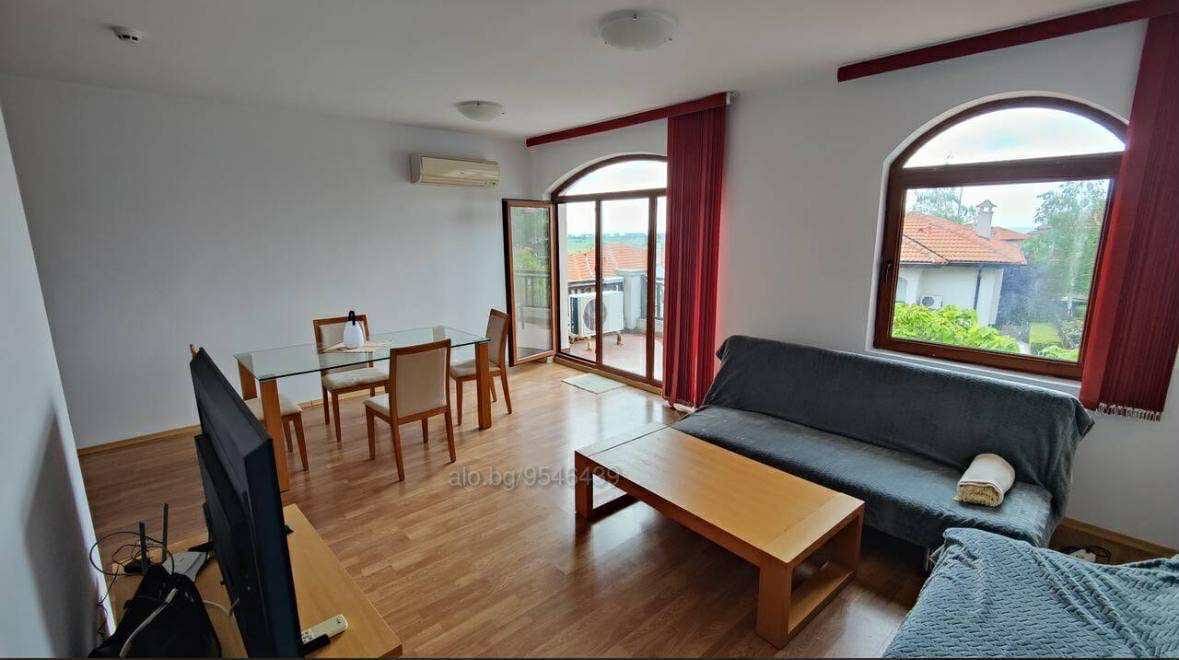 Светлая гостиная с большим окном, мебелью и выходом на балкон, идеальная для покупки недвижимости в Болгарии.