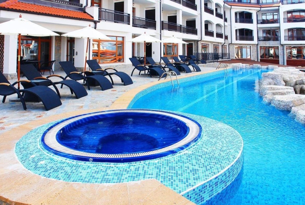 Современный бассейн с джакузи в комплексе недвижимости в Болгарии, окружённый шезлонгами и зонтиками для отдыха.