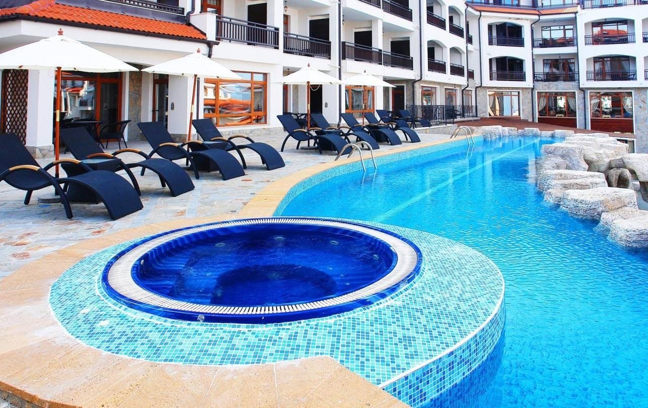 Современный бассейн с джакузи в комплексе недвижимости в Болгарии, окружённый шезлонгами и зонтиками для отдыха.