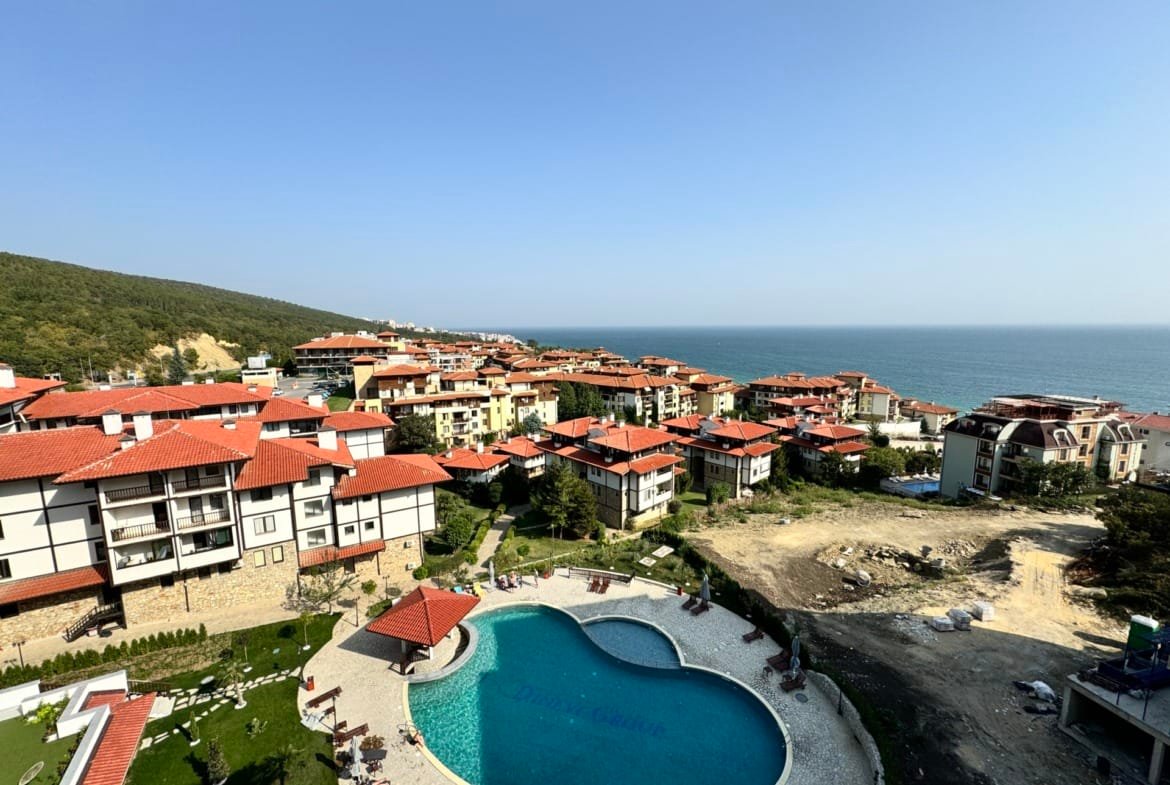 Жилой комплекс с бассейном и видом на море в Болгарии, окружённый зелеными холмами и зданиями в традиционном стиле.