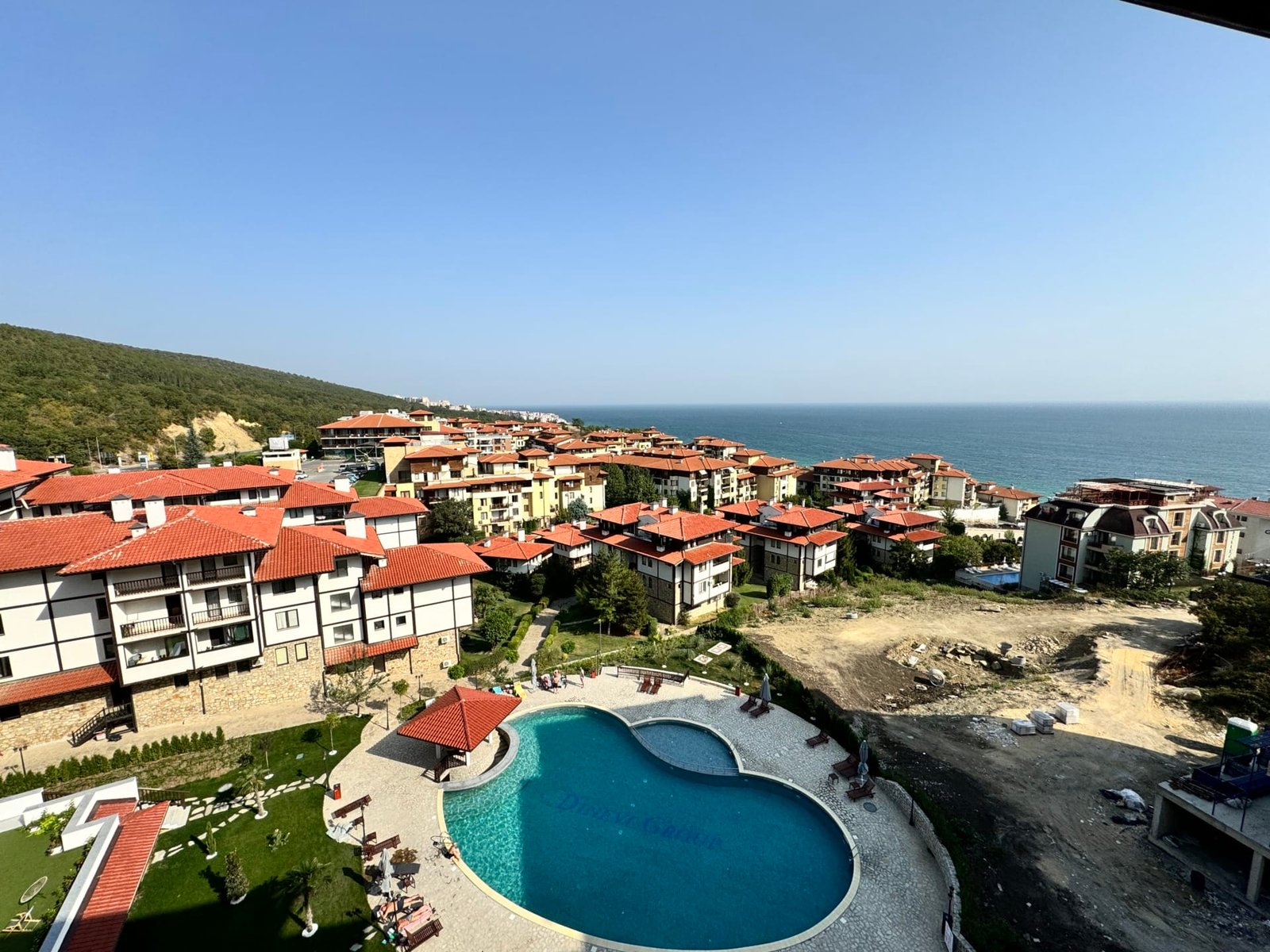 Жилой комплекс с бассейном и видом на море в Болгарии, окружённый зелеными холмами и зданиями в традиционном стиле.