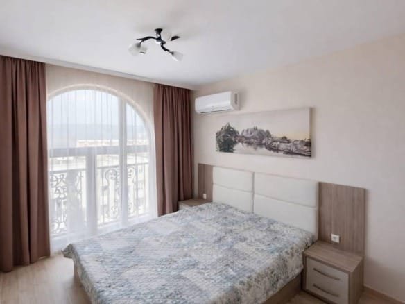 Уютная спальня с балконом, мягким освещением, светлыми тонами и современными мебельными решениями для покупки недвижимости в Болгарии.