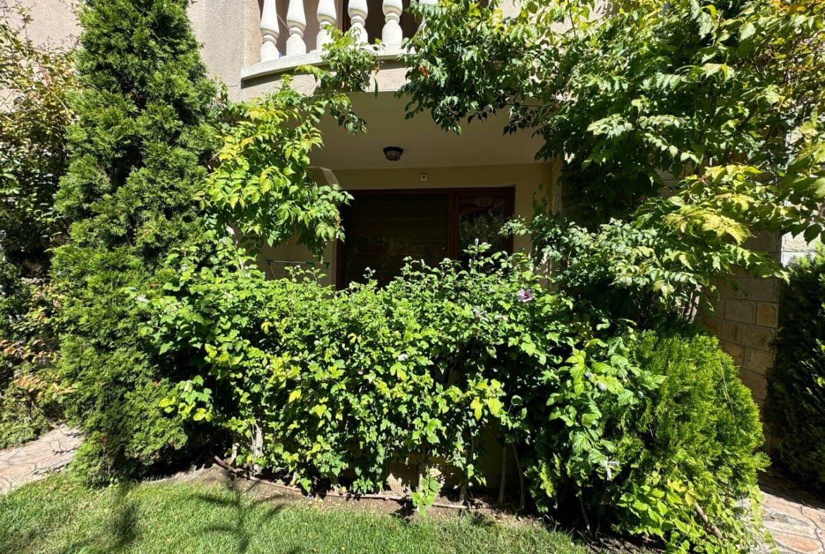 Уютный вход в дом, окруженный зелеными кустарниками и деревьями, на фоне солнечного дня в Болгарии. Оптимально для покупки недвижимости.